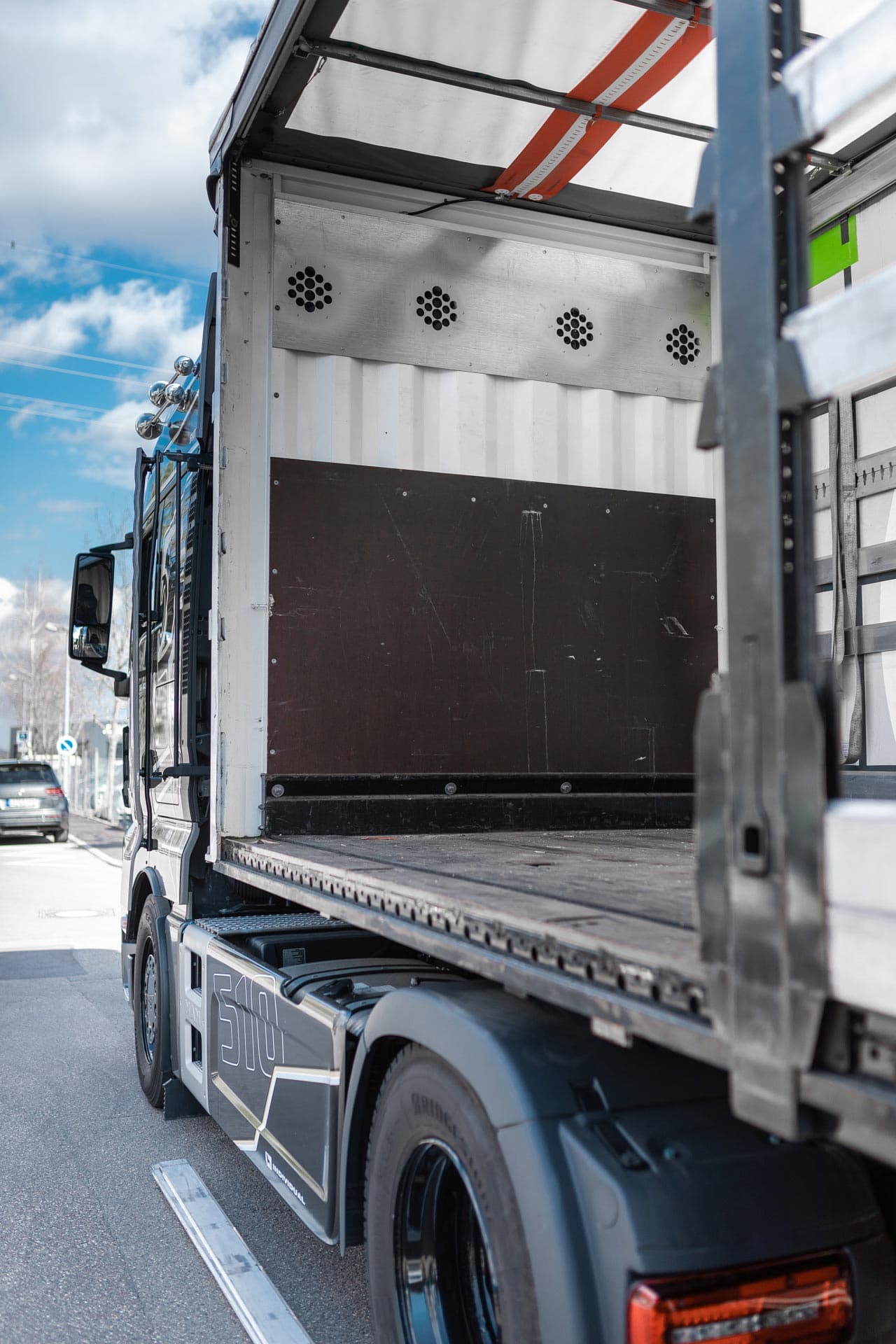 Un camion cu suprastructura argintie și anvelope negre, parcat sub cerul albastru.
