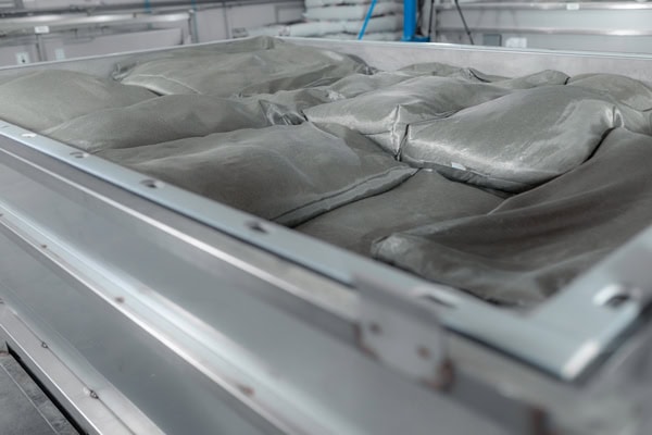 O cadă industrială din oțel inoxidabil, de culoare gri, plină cu saci nedeschisi conținând pulbere gri, redată în detaliu.
