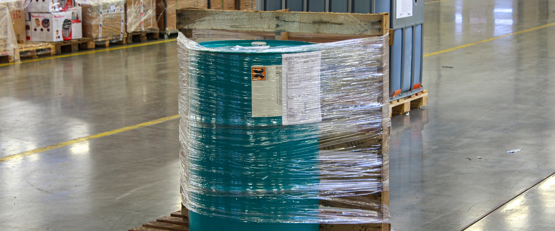 Un butoi verde, învelit în plastic și fixat pe o paletă de lemn. Butoiul este înconjurat de mărfuri ambalate și de un alt container mare, ceea ce sugerează un mediu de depozitare bine organizat.